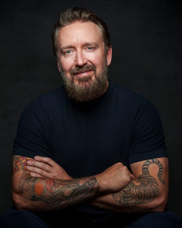 Chad Daniels in a black T-shirt against a charcoal background, arms crossed, both forearms tattooed.