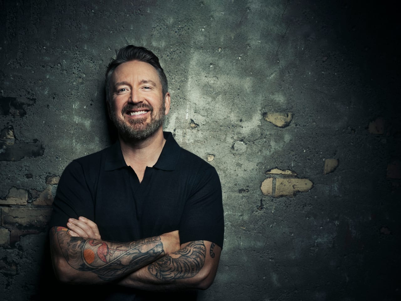 Chad Daniels against a broken concrete wall, arms crossed, laughing.