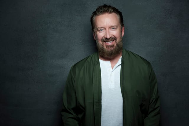 Chad Daniels in an olive bomber jacket against a charcoal background.