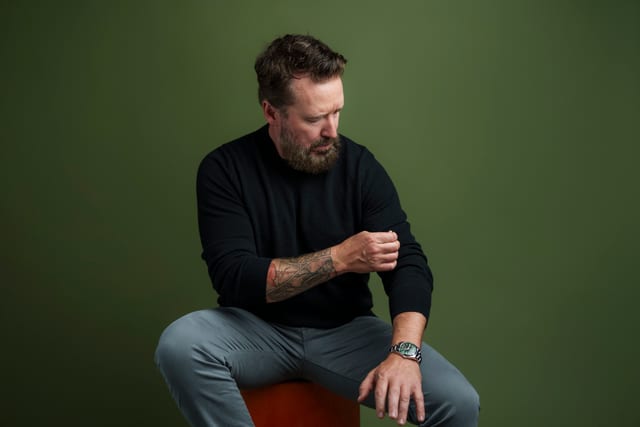 Chad Daniels seated against a green background, looking down.