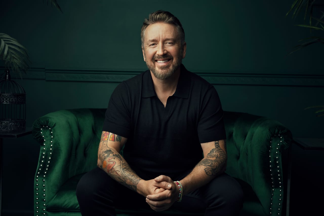 Chad Daniels seated on a green velvet Chesterfield in a dark panelled room, hands clasped, both forearms tattooed.