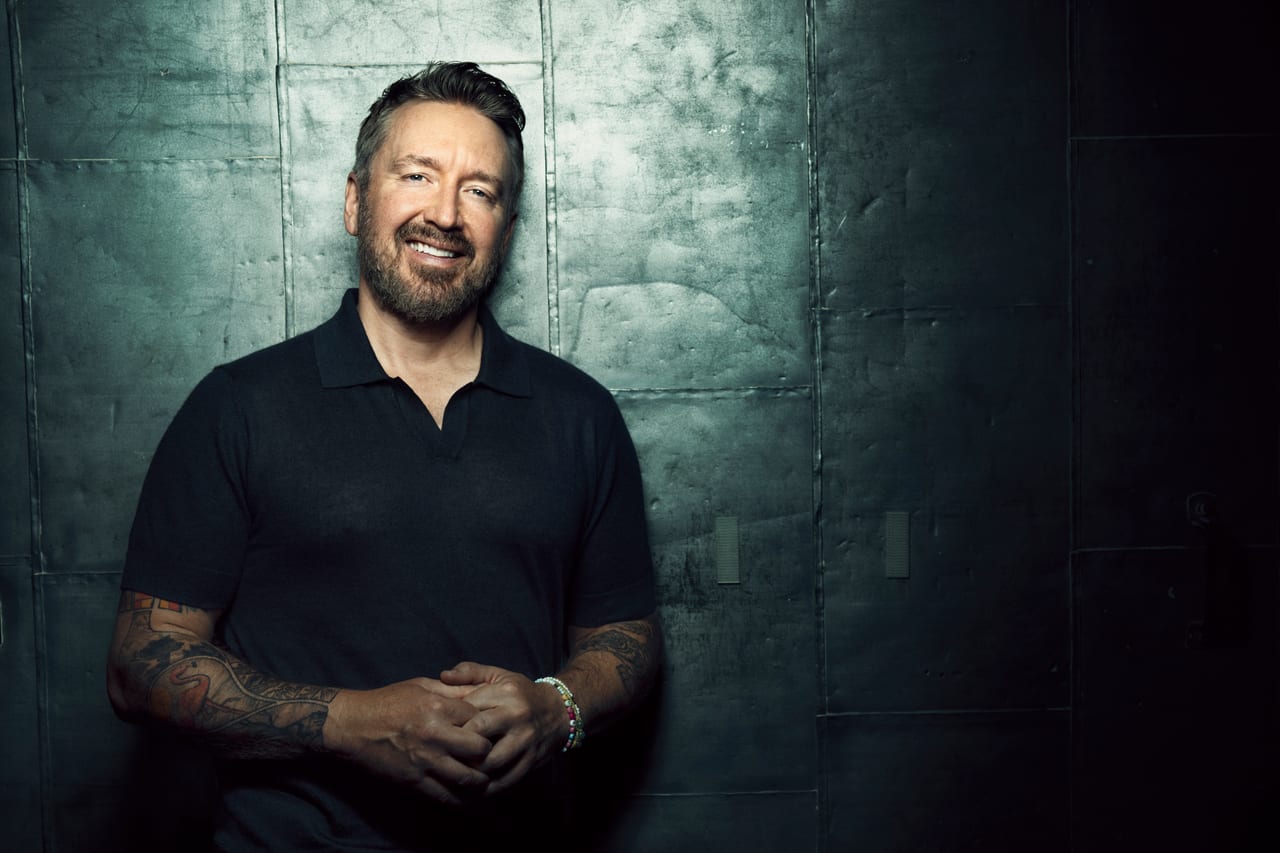 Chad Daniels seated against a grey panelled wall, hands clasped, both forearms tattooed.