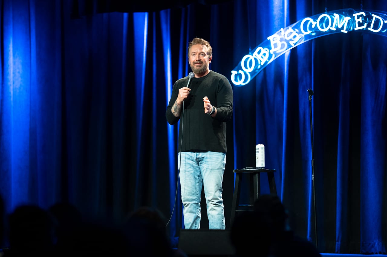 Chad Daniels on stage at Cobb's Comedy Club, microphone in hand, the club's neon sign lit behind him and the room in silhouette.