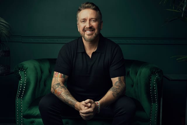 Chad Daniels seated on a green velvet Chesterfield, hands clasped.