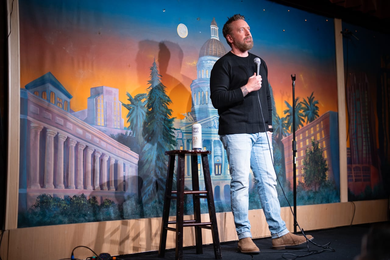 Chad Daniels on stage in front of a painted mural of the Sacramento capitol at sunset, microphone in hand.