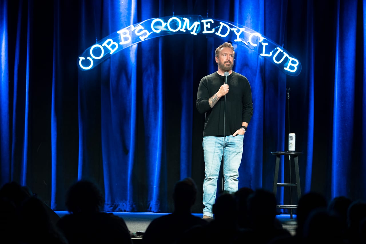 Chad Daniels on stage at Cobb's Comedy Club under the club's blue neon sign, the room dark in front of him.